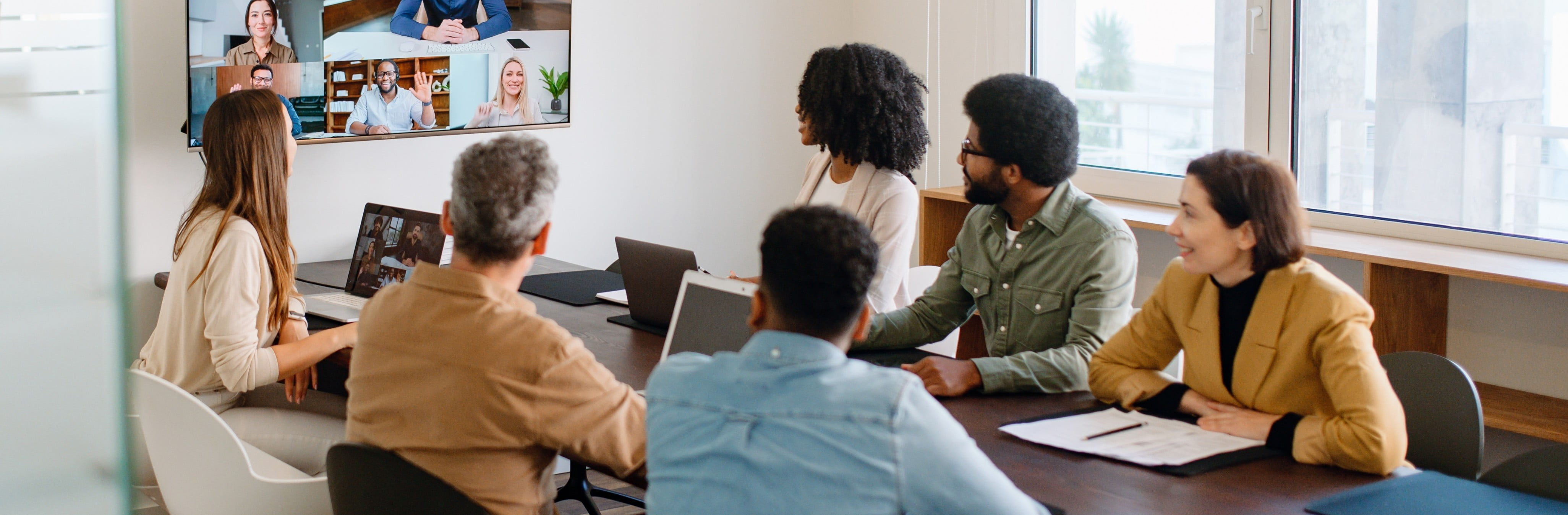 A board room meeting with a bunch of people sitting at a board table and a big screen with more people on a video call.